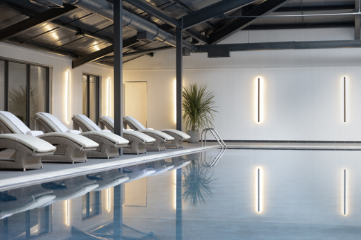 Indoor luxury swimming pool at Mar Hall Resort Glasgow with turquoise water, surrounded by elegant stone tiling, white Roman-style pillars, and large windows.
